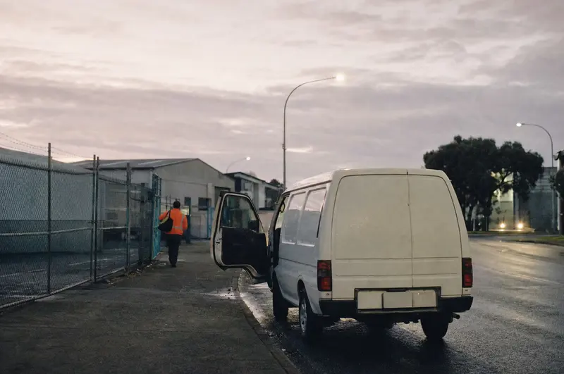 A white work van with its door open at dawn on an industrial street in Wiri, South Auckland, a worker in hi-vis walking to the warehouse gate
