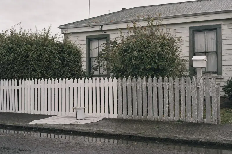 Half-repainted white picket fence outside a South Dunedin cottage on a grey morning, a paint tin on the footpath