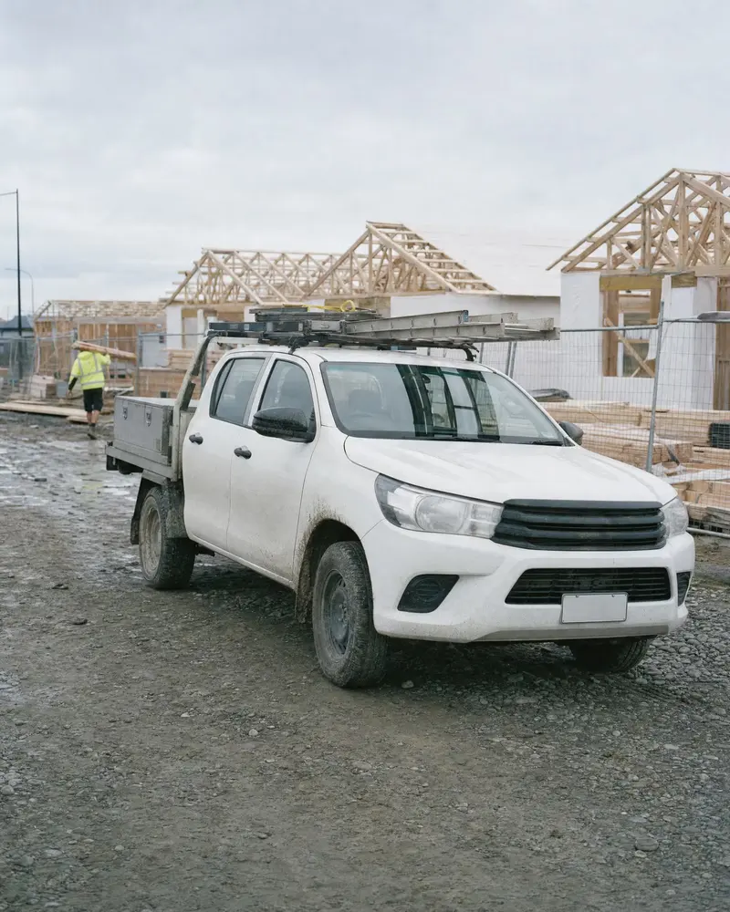 A white work ute with a ladder on the rack and a toolbox on the deck at a new subdivision in Rolleston, Canterbury, a builder carrying timber in the distance