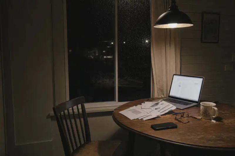 Debt consolidation paperwork on a kitchen table at night: a laptop, a pile of bills and a cold cup of tea, rain on the window