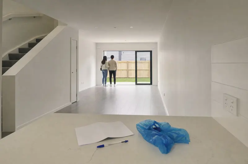 A Saturday open home at a new townhouse in Papakura, Auckland: a folded listing sheet and shoe covers on the kitchen bench while a couple look out at the courtyard