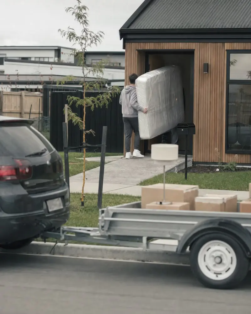 Moving day at a first home in Wigram, Christchurch: a mattress going through the front door of a new cedar-clad townhouse, a borrowed trailer of boxes at the kerb