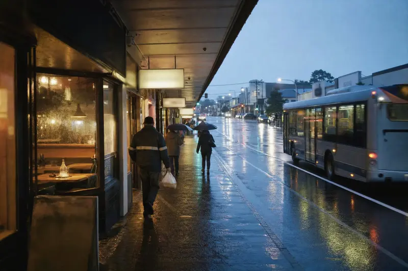 People walking under the shop verandas on Dominion Road, Auckland, at dusk in light rain as a bus pulls away