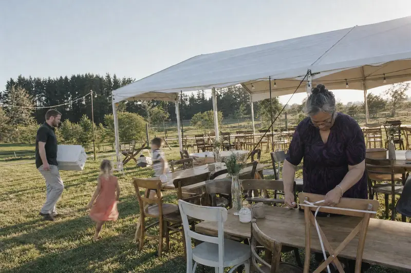 Family sets up a backyard wedding marquee on a Waikato lifestyle block, an aunty tying ribbon to borrowed chairs